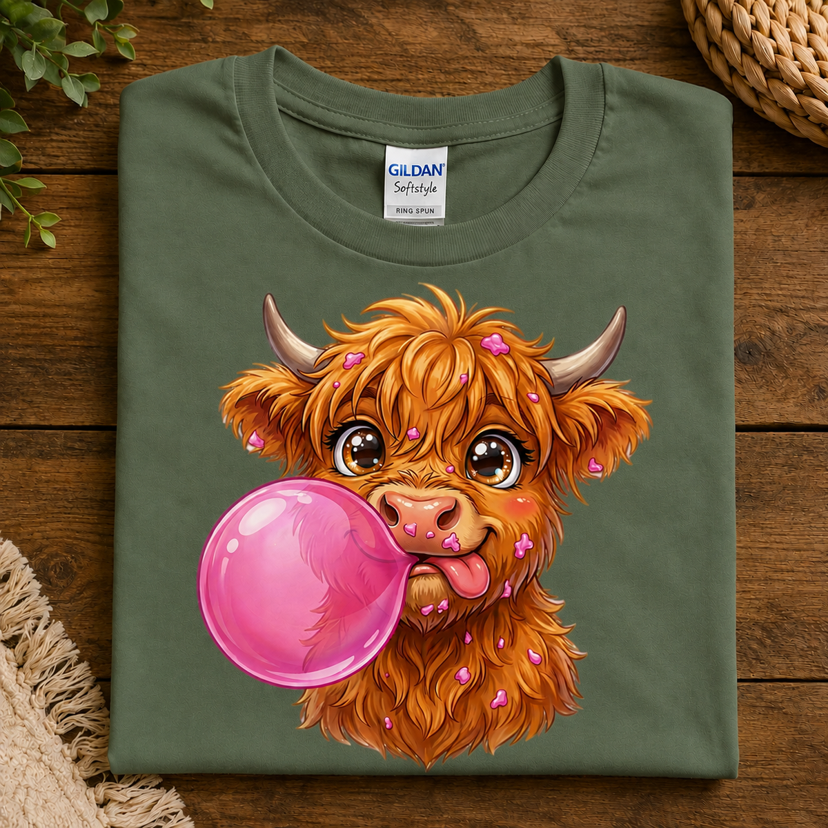A folded Military green Gildan Softstyle T-shirt with a colorful cartoon illustration of a fluffy orange Highland cow blowing a large pink bubblegum bubble, displayed on a rustic wooden surface.