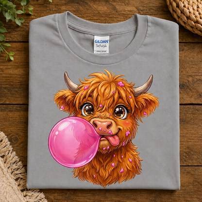 A folded sport gray Gildan Softstyle T-shirt with a colorful cartoon illustration of a fluffy orange Highland cow blowing a large pink bubblegum bubble, displayed on a rustic wooden surface.