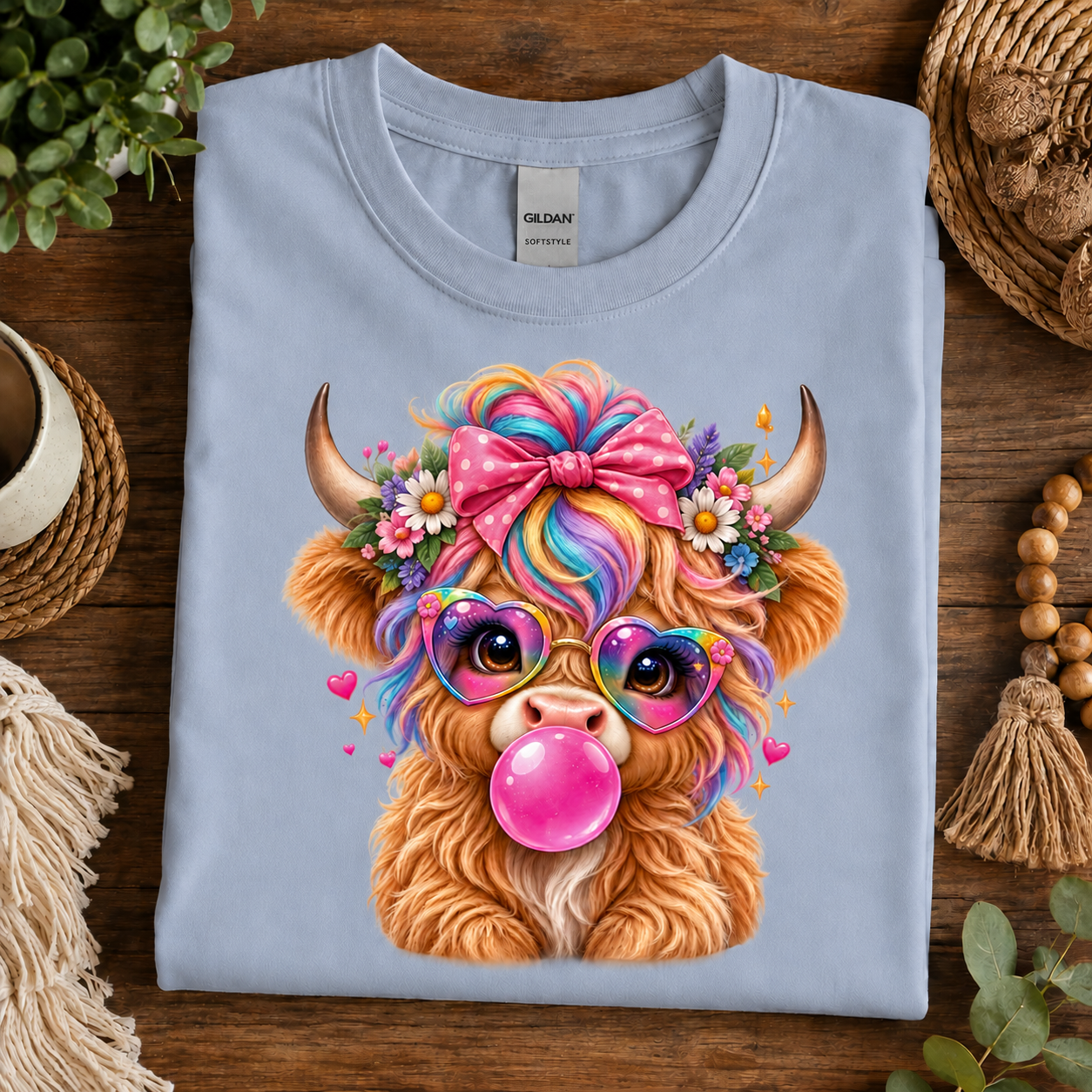 A folded light blue Gildan t-shirt with a colorful cartoon highland cow graphic, displayed on a dark wooden table surrounded by lifestyle props like a plant, woven basket, and beads.