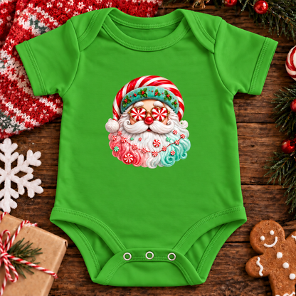 A apple green baby bodysuit with a colorful Christmas Santa graphic on the chest, styled in a flat-lay on a rustic wooden surface with holiday decorations including a gingerbread cookie, pine branches, a snowflake, and a wrapped gift.