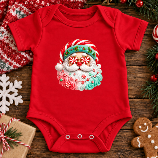 A red baby bodysuit with a festive Christmas Santa graphic, laid flat on a rustic wooden surface surrounded by holiday props including a gingerbread cookie, pine branches, a gift box, and a white snowflake.