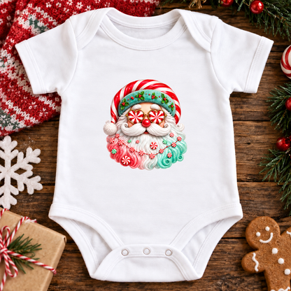 A white baby bodysuit with a colorful Christmas Santa graphic, laid flat on a rustic wooden surface surrounded by holiday decorations including a red knit blanket, pine branches, a gingerbread cookie, and a wrapped gift.