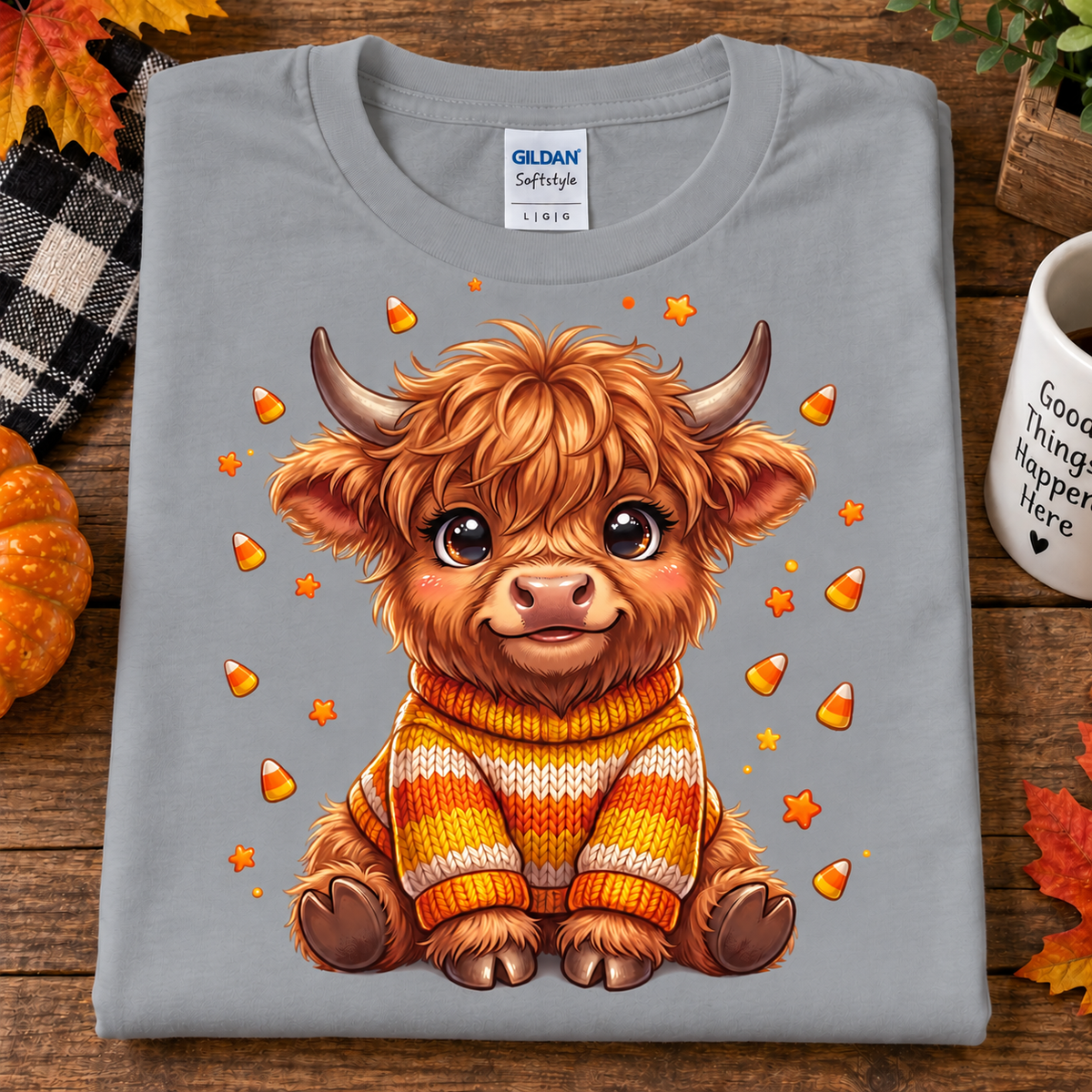 A folded sport gray Gildan Softstyle t-shirt is displayed on a rustic wooden surface. The shirt features a detailed graphic of a cute, fluffy Highland cow wearing a yellow, orange, and white striped knit sweater, surrounded by scattered candy corn and orange stars.