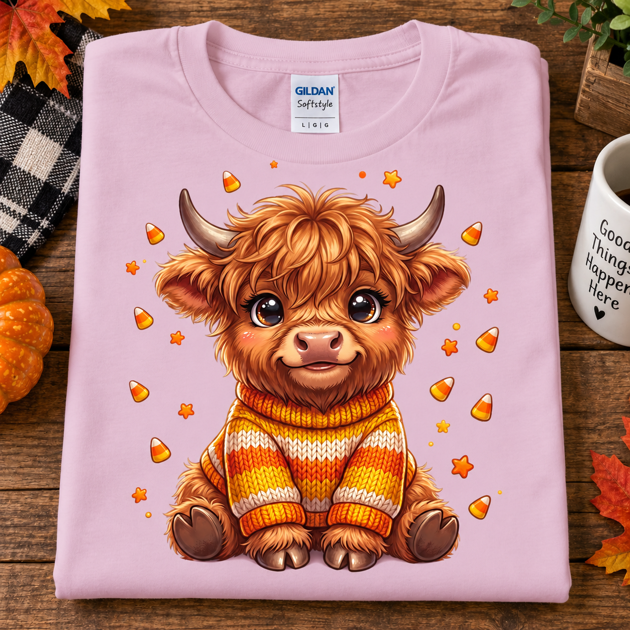 A folded light pink Gildan Softstyle Linen T-shirt is displayed on a rustic wooden surface. The shirt features a cute cartoon illustration of a fluffy brown highland cow wearing a yellow, orange, and white striped knit sweater, surrounded by candy corn and small orange stars.