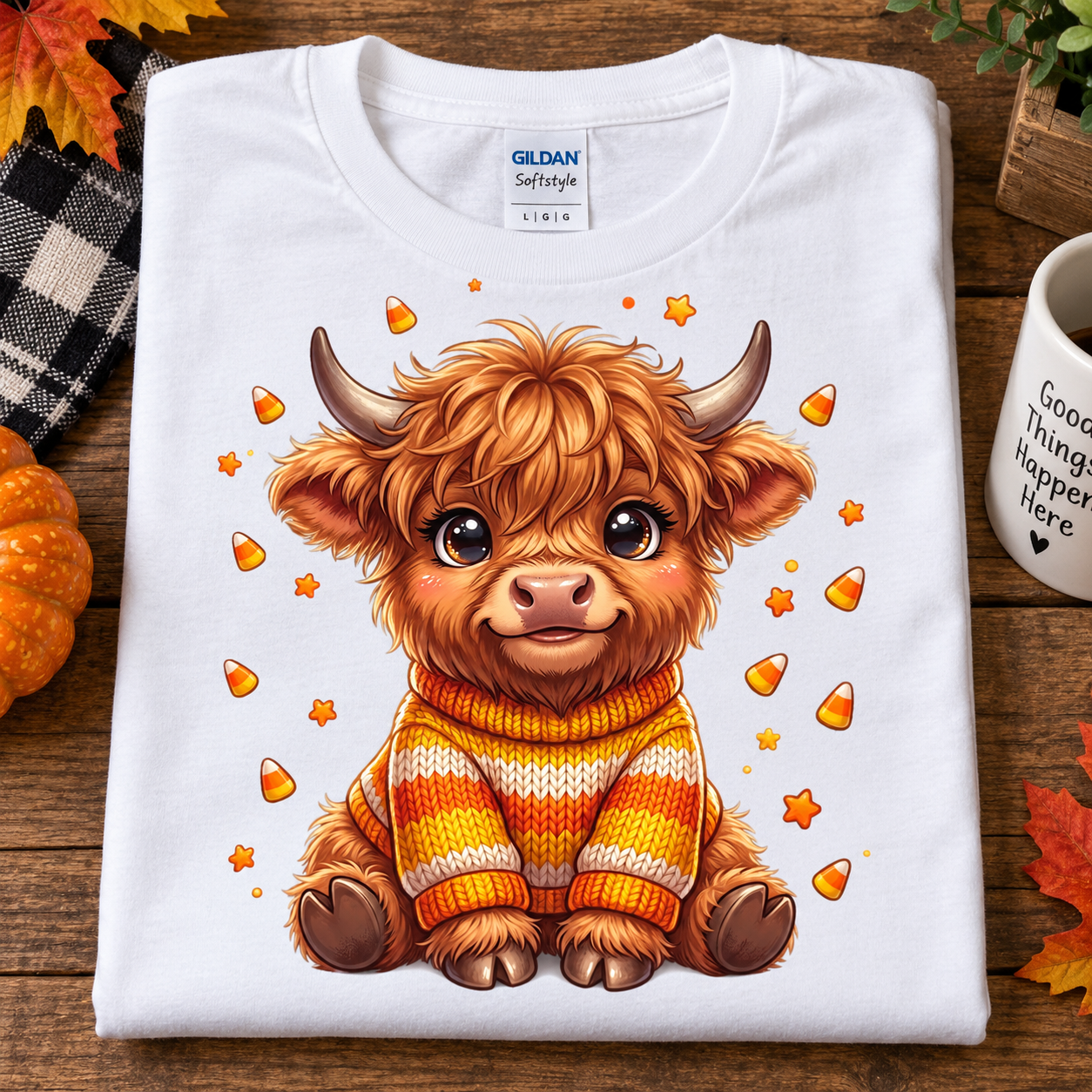 A folded white Gildan Softstyle T-shirt with a cartoon illustration of a fluffy brown Highland cow wearing a yellow, orange, and white striped sweater, surrounded by candy corn and stars. The shirt is styled on a wooden surface with autumn leaves, a pumpkin, a plaid blanket, and a mug.