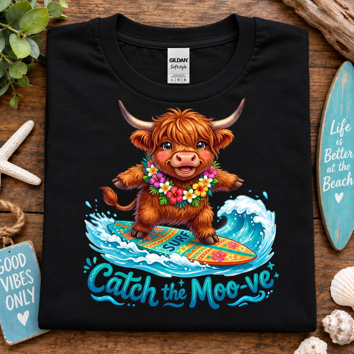 A black Gildan Straightforward t-shirt laid flat on a rustic wooden surface, featuring a colorful graphic of a fluffy brown Highland cow surfing on a wave with the text 'Catch the Moo-ve'. The shirt is styled with beach-themed props including a starfish, seashells, a turquoise surfboard, and a small sign.