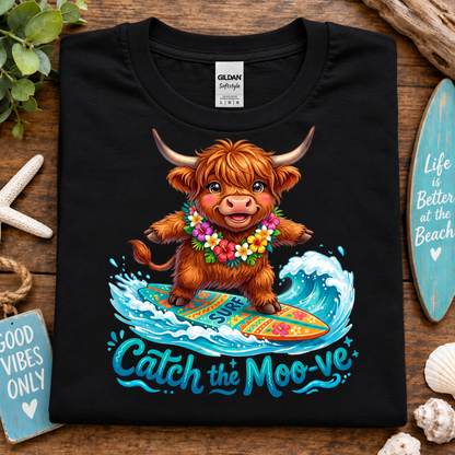 A black Gildan Straightforward t-shirt laid flat on a rustic wooden surface, featuring a colorful graphic of a fluffy brown Highland cow surfing on a wave with the text 'Catch the Moo-ve'. The shirt is styled with beach-themed props including a starfish, seashells, a turquoise surfboard, and a small sign.