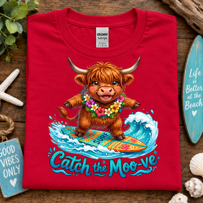 A folded red Gildan Satinway t-shirt with a colorful graphic of a cartoon Highland cow surfing on a wave with the text 'Catch the Moo-ve'. The shirt is displayed on a rustic wooden background surrounded by beach-themed props.