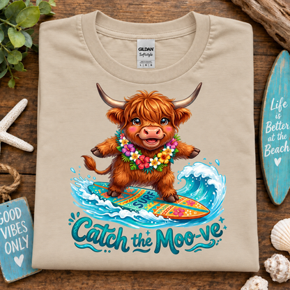 A sand colored Gildan Signature t-shirt laid flat on a rustic wooden surface, featuring a colorful graphic of a Highland cow surfing with the text 'Catch the Moo-ve'. The shirt is styled with beach-themed props including a starfish, shells, a wooden sign, and driftwood.