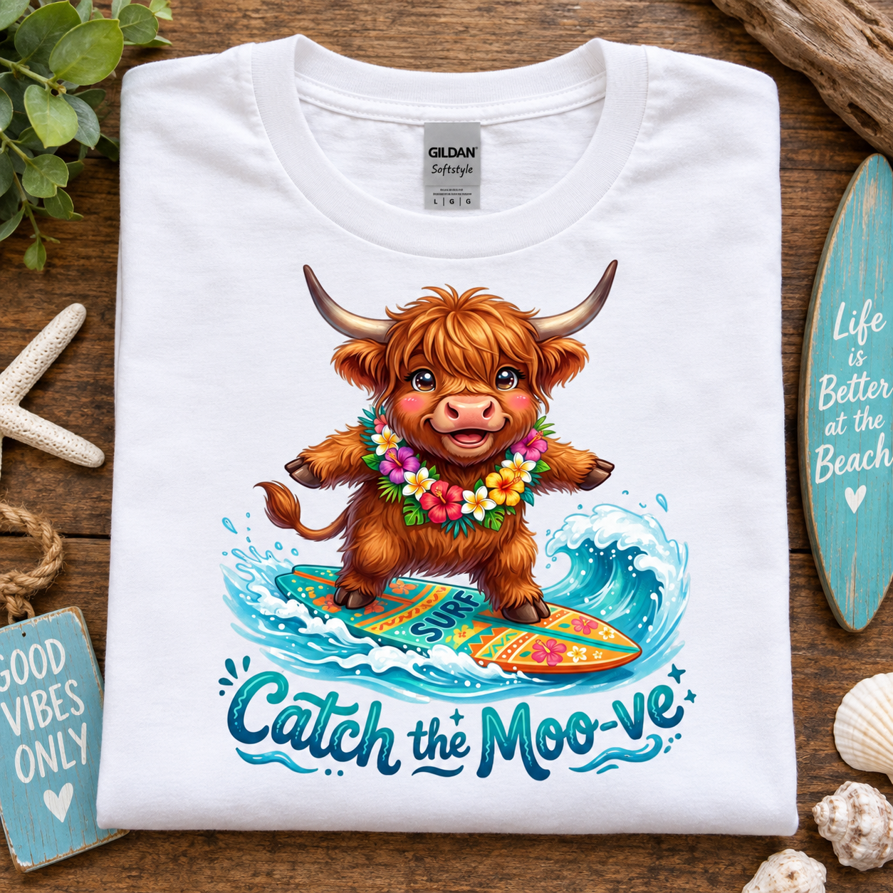 A white Gildan Straightforward t-shirt laid flat on a rustic wooden surface, featuring a colorful graphic of a fluffy brown Highland cow surfing on a wave with the text 'Catch the Moo-ve'. The shirt is styled with beach-themed props including a starfish, seashells, a turquoise surfboard, and a small sign.