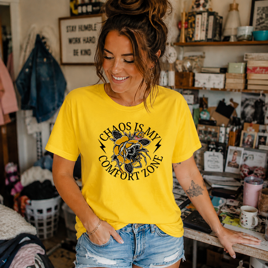 A woman wearing a bright yellow graphic t-shirt with a black circular design that reads 'CHAOS IS MY COMFORT ZONE' around a central floral motif, paired with denim shorts in a cluttered boutique interior.