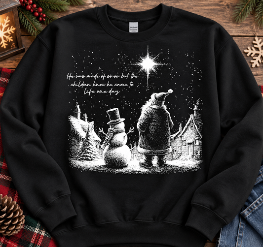 A black crewneck sweatshirt featuring a white graphic print of Santa Claus, a snowman, and a cozy village under a bright starry night sky. The text on the chest reads, 'He was made of snow but the children know he came to life once more.