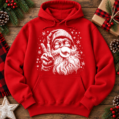 A red pullover hoodie with a white graphic of a Santa Claus figure blowing a bubblegum bubble, styled in a flat lay on a rustic wooden surface with holiday decorations like pine branches, a gift box, and a star.