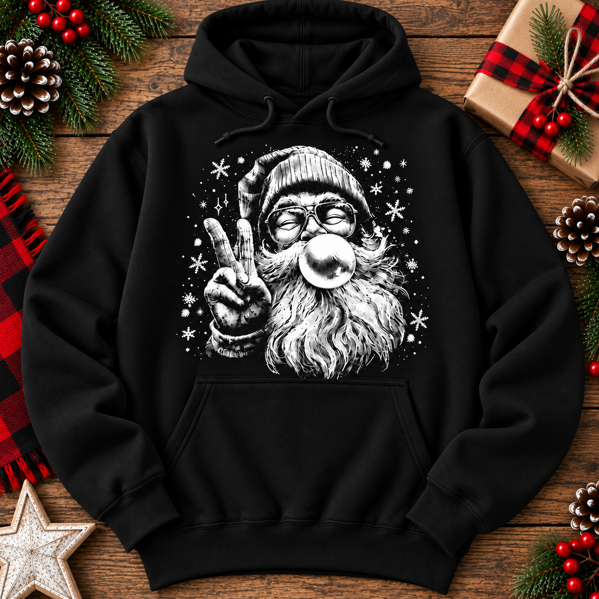 A black pullover hoodie laid flat on a rustic wooden surface, decorated with Christmas-themed props including pinecones, red berries, a wrapped gift, and a star. The hoodie features a large white graphic print of Santa Claus blowing a bubblegum bubble and making a peace sign.