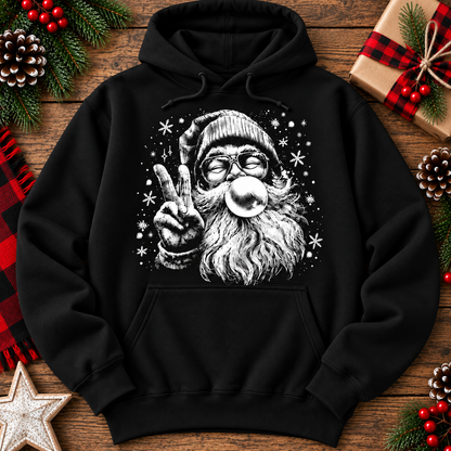 A black pullover hoodie laid flat on a rustic wooden surface, decorated with Christmas-themed props including pinecones, red berries, a wrapped gift, and a star. The hoodie features a large white graphic print of Santa Claus blowing a bubblegum bubble and making a peace sign.