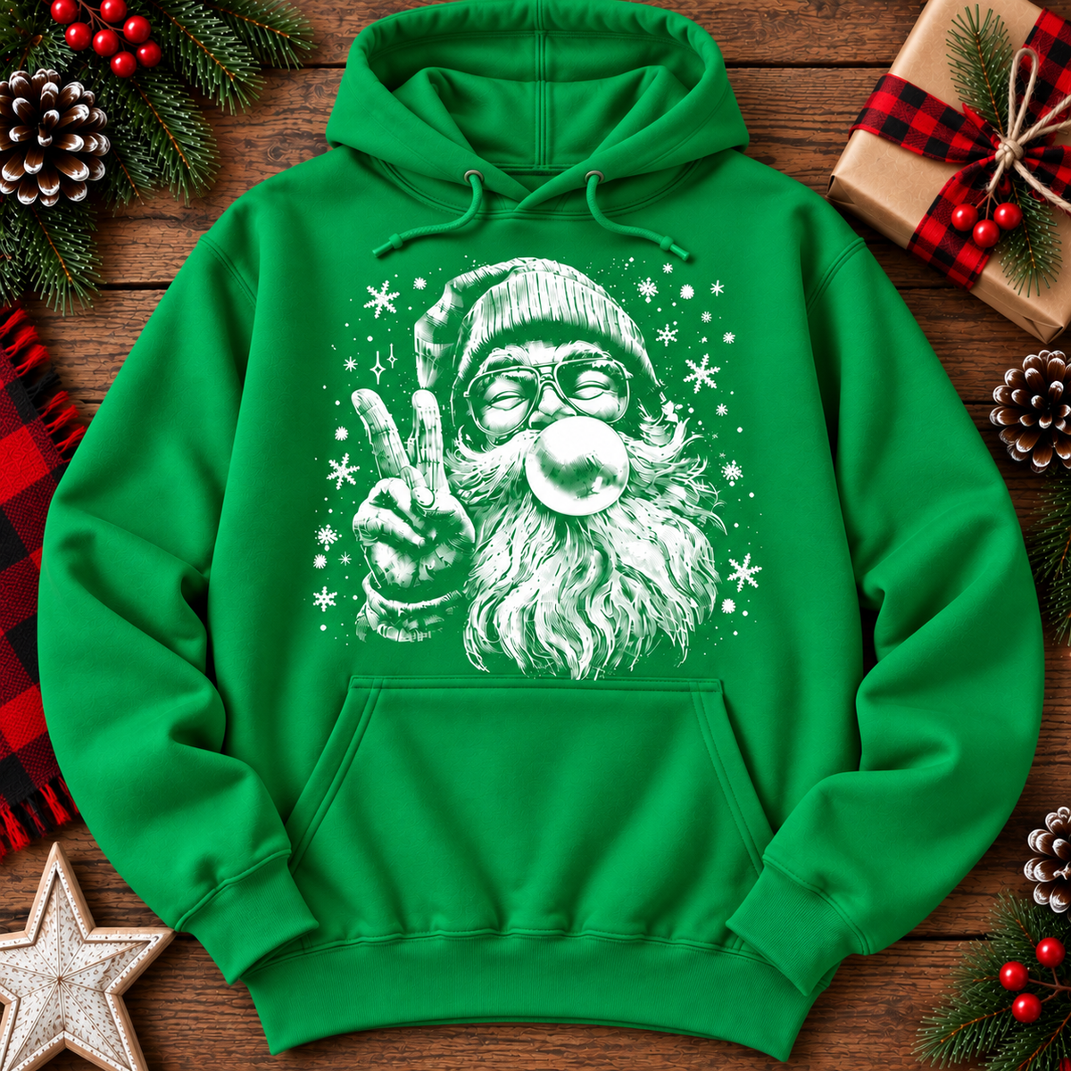 A bright green pullover hoodie laid flat on a rustic wooden surface, featuring a large white graphic of Santa Claus wearing glasses and making a peace sign while blowing a bubble. The hoodie is surrounded by holiday decorations including pine cones, red berries, a wrapped gift, and a star.