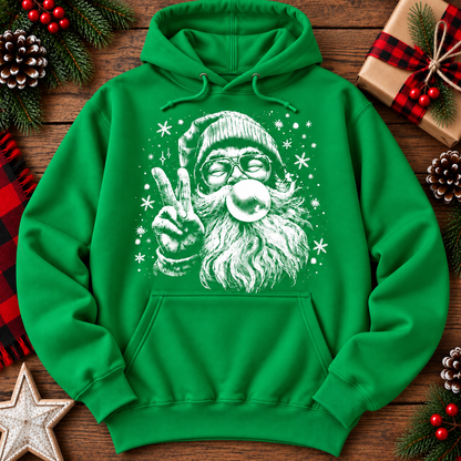 A bright green pullover hoodie laid flat on a rustic wooden surface, featuring a large white graphic of Santa Claus wearing glasses and making a peace sign while blowing a bubble. The hoodie is surrounded by holiday decorations including pine cones, red berries, a wrapped gift, and a star.