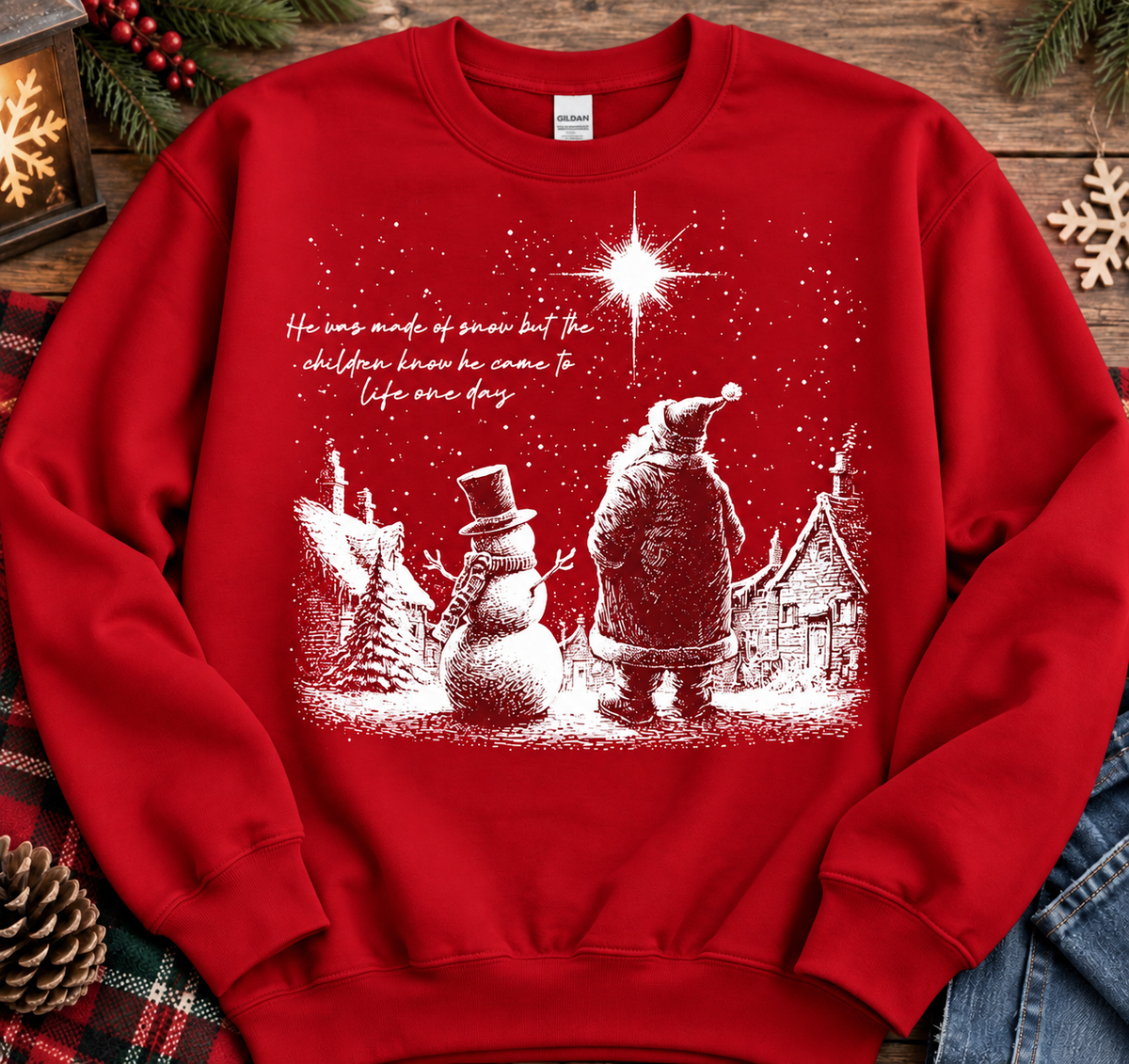 A red crewneck sweatshirt with a white Christmas graphic print of Santa Claus, a snowman, a star, and a cozy house, styled flat on a rustic wooden background with holiday decorations.