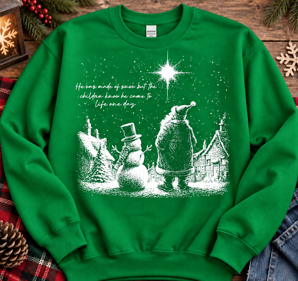 A green crewneck sweatshirt with a white Christmas graphic of Santa Claus, a snowman, and a starry night sky, laid flat on a rustic wooden background with holiday decorations.