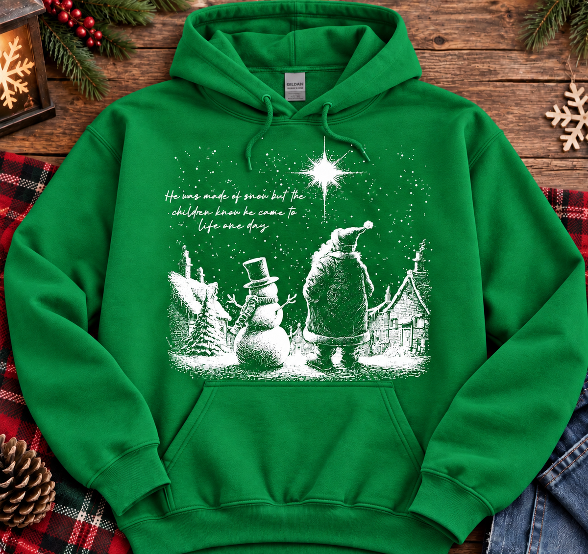 A bright green pullover hoodie with a white Christmas-themed graphic print on the front, laid flat on a rustic wooden surface with holiday decorations and a plaid blanket around it.