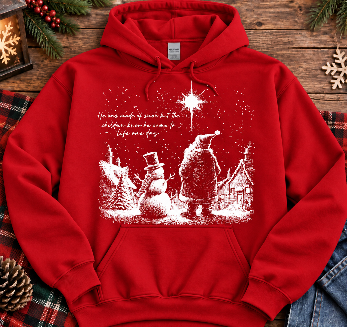 A red pullover hoodie with a white Christmas-themed graphic print of Santa Claus, a snowman, and houses under a star, laid flat on a rustic wooden background with holiday decorations.