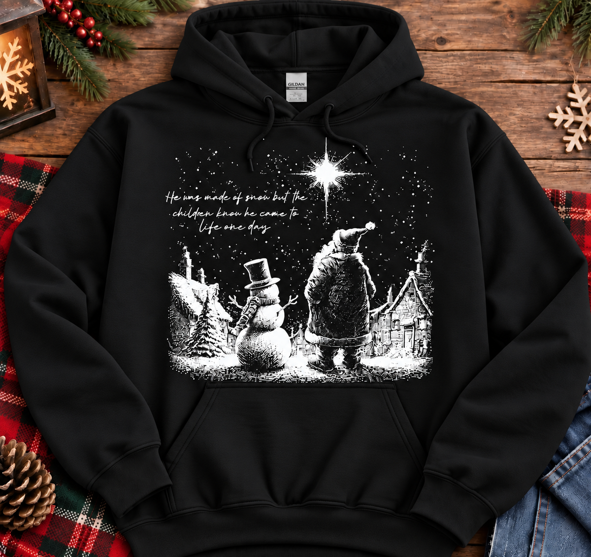 A black pullover hoodie laid flat on a rustic wooden surface, decorated with holiday props including pine branches, a lantern, red plaid fabric, and jeans. The hoodie features a white Christmas village graphic with Santa, a snowman, a house, and a bright star, plus small cursive text on the chest.