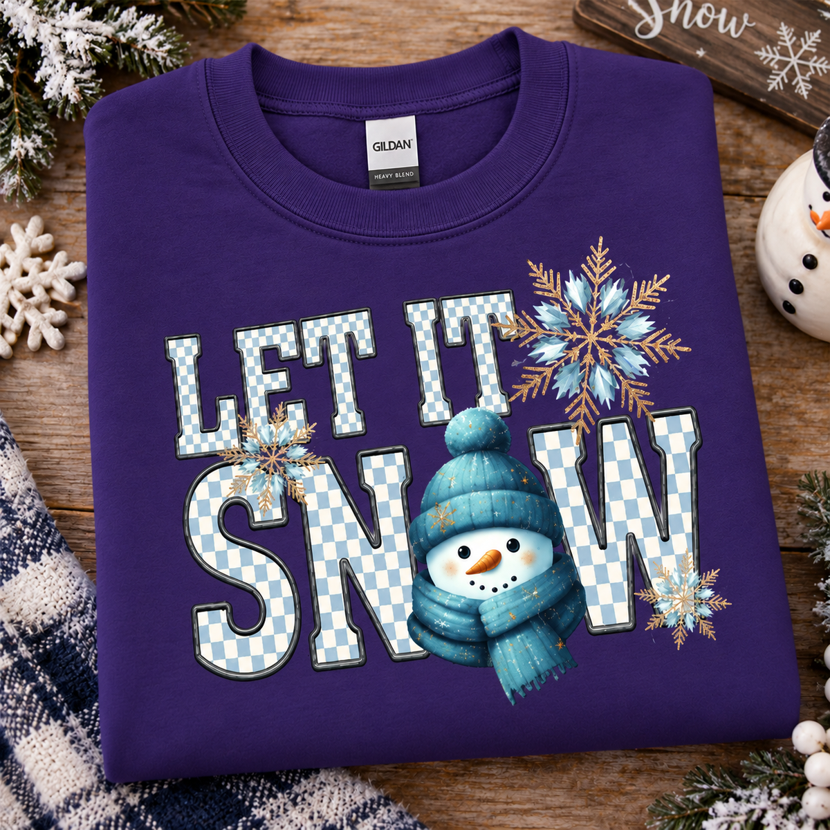 A folded purple Gildan sweatshirt with a 'LET IT SNOW' graphic featuring a snowman and snowflakes, displayed on a rustic wooden background with holiday decorations.