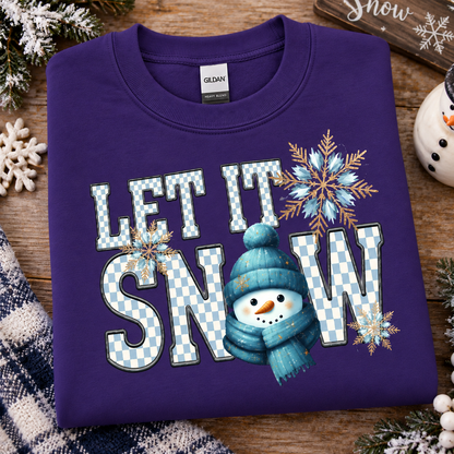 A folded purple Gildan sweatshirt with a 'LET IT SNOW' graphic featuring a snowman and snowflakes, displayed on a rustic wooden background with holiday decorations.