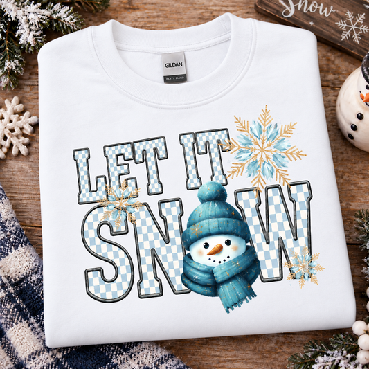 A folded white Gildan sweatshirt with a 'LET IT SNOW' graphic featuring a blue snowman, blue and white checkered lettering, and gold snowflakes, styled on a wooden surface with holiday decorations.