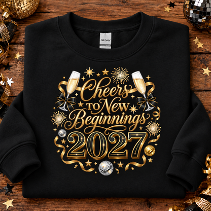 A black crewneck sweatshirt with a gold and silver graphic reading 'Cheers to New Beginnings 2027' surrounded by champagne glasses, stars, and fireworks, laid flat on a rustic wooden background with holiday decorations.