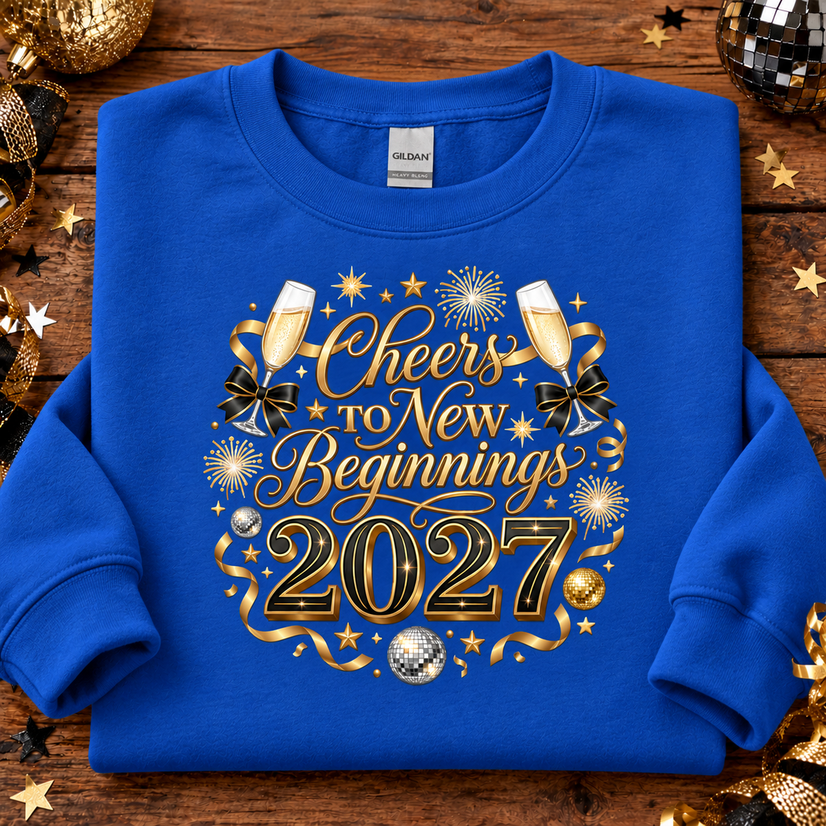 A sapphire blue crewneck sweatshirt with a festive 'Cheers to New Beginnings 2027' graphic, laid flat on a rustic wooden background decorated with gold and silver holiday ornaments.