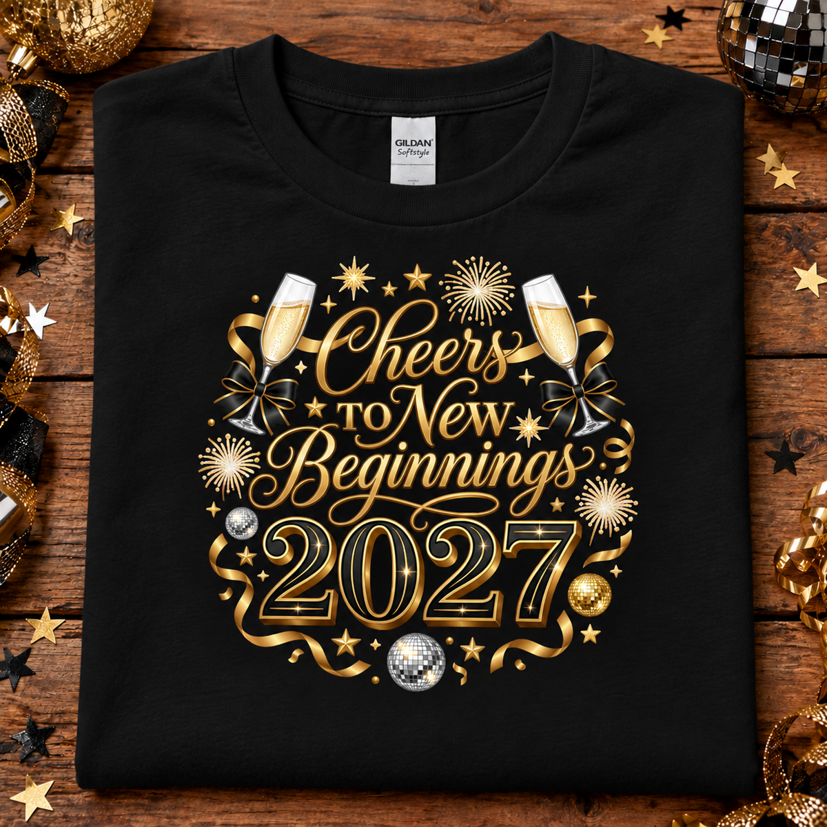 A black Gildan t-shirt with a gold and silver graphic reading 'Cheers to New Beginnings 2027' surrounded by champagne glasses, stars, and fireworks, laid flat on a rustic wooden background with holiday decorations.