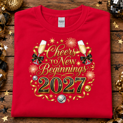A folded red Gildan Comfort T-shirt with a festive 'Cheers to New Beginnings 2027' graphic, displayed on a rustic wooden background decorated with gold and black holiday ornaments.