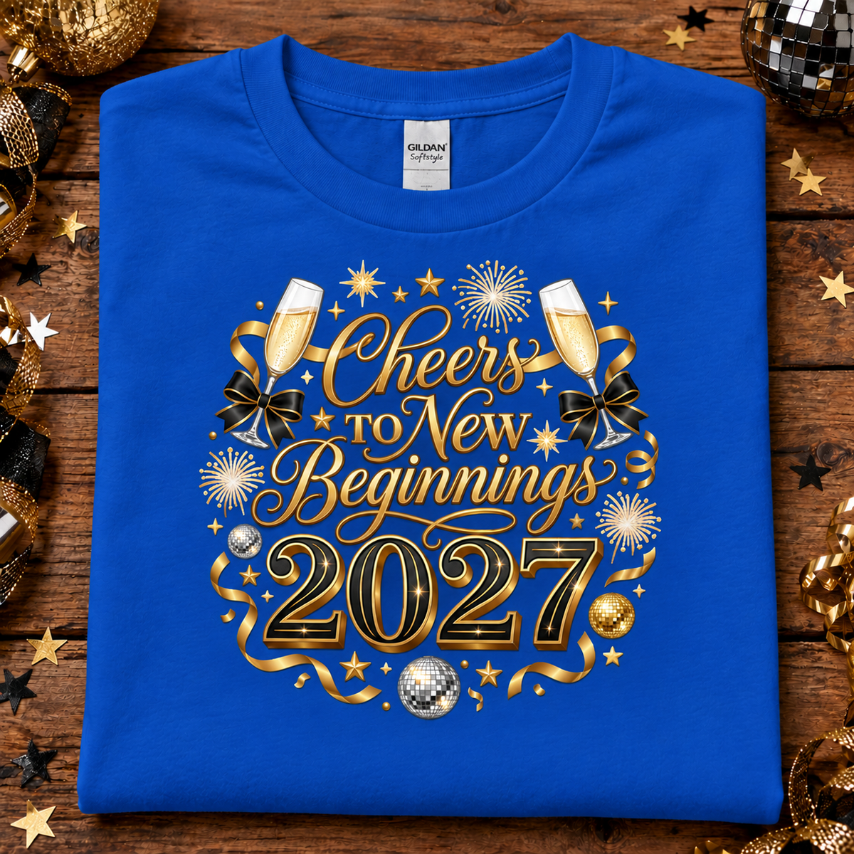 A folded sapphire blue Gildan Softstyle t-shirt with a 'Cheers to New Beginnings 2027' graphic, displayed on a rustic wooden background decorated with gold and black party ornaments.