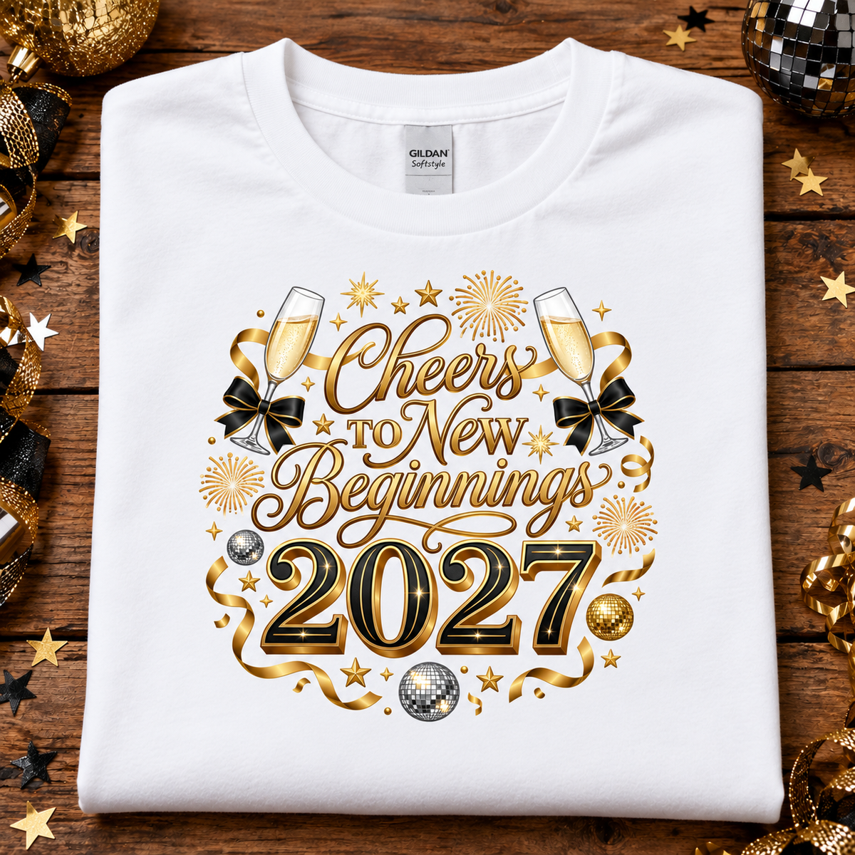 A white Gildan t-shirt laid flat on a rustic wooden background, decorated with gold and black New Year's Eve themed graphics that read 'Cheers to New Beginnings 2027' with champagne glasses, bows, stars, and fireworks. Gold and black party decorations are scattered around the edges.