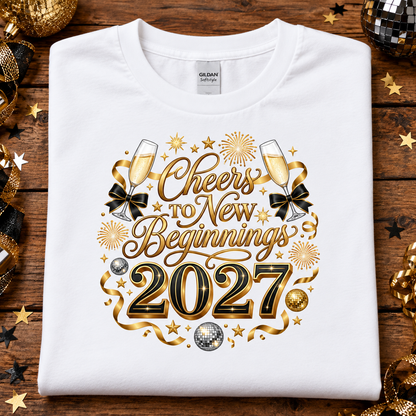 A white Gildan t-shirt laid flat on a rustic wooden background, decorated with gold and black New Year's Eve themed graphics that read 'Cheers to New Beginnings 2027' with champagne glasses, bows, stars, and fireworks. Gold and black party decorations are scattered around the edges.
