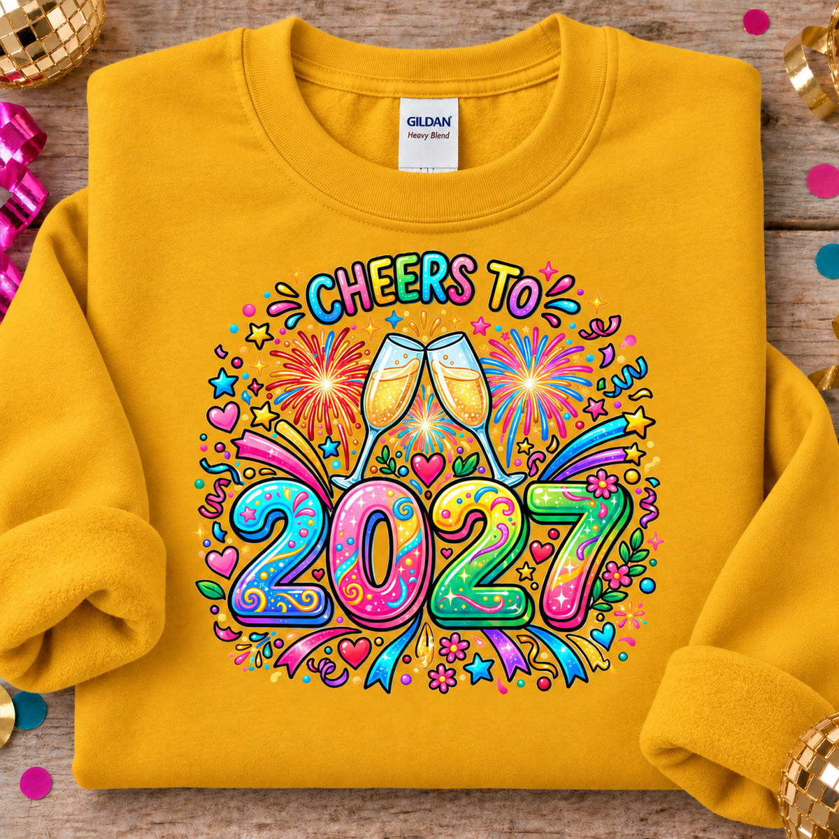 A folded gold colored Gildan Heavy Blend sweatshirt on a rustic wooden background, featuring a colorful 'Cheers to 2027' graphic with champagne glasses, fireworks, and confetti.