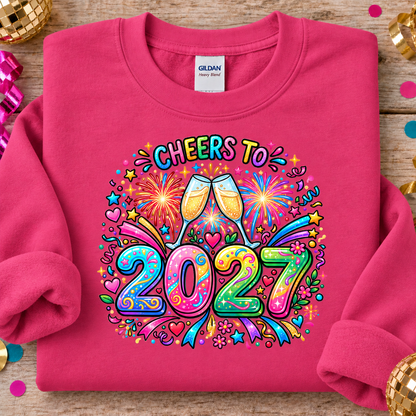 A folded heliconia pink crewneck sweatshirt on a rustic wooden surface, decorated with party props like a disco ball and confetti. The sweatshirt features a colorful graphic print that reads 'CHEERS TO 2027' with two clinking champagne glasses, fireworks, and rainbow accents.