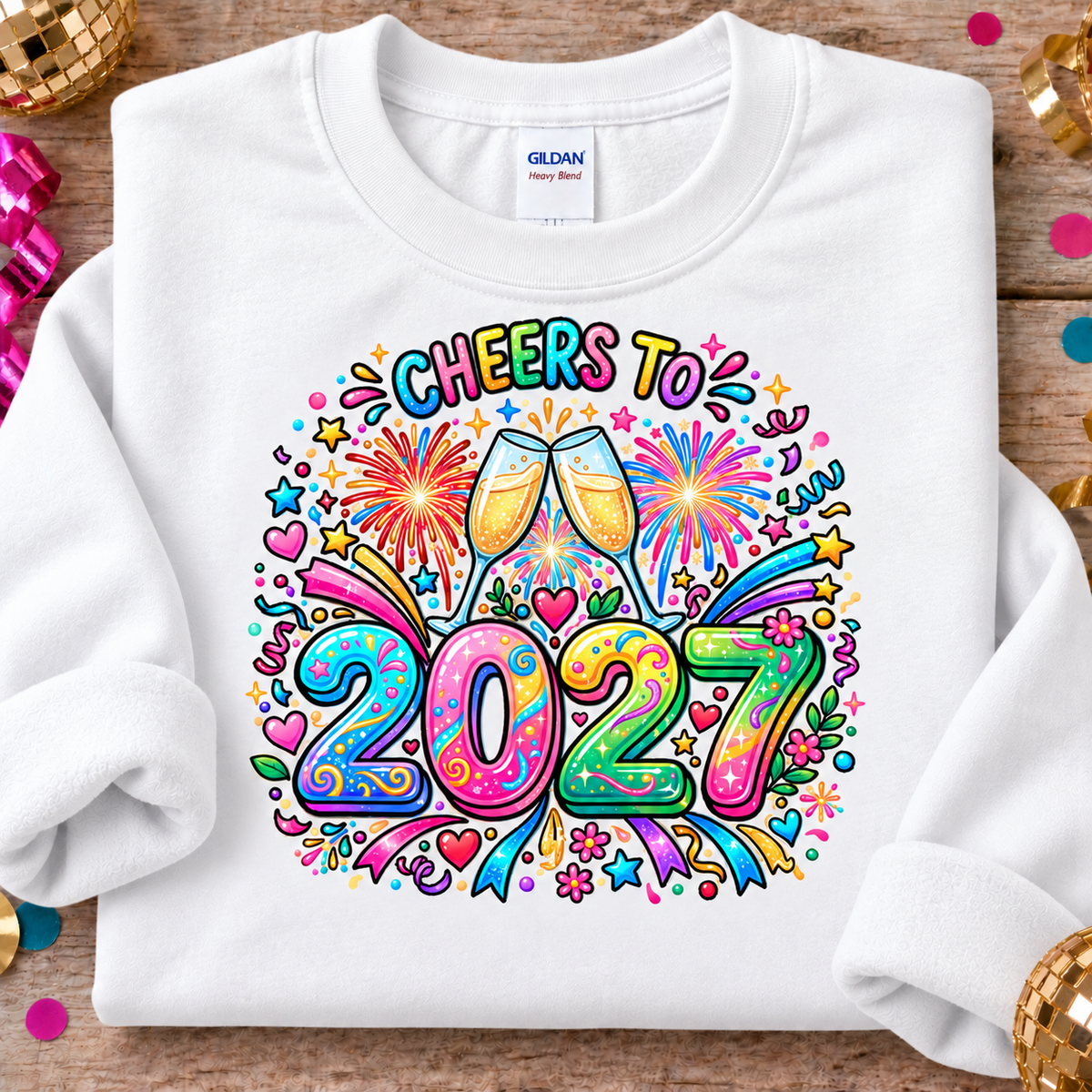 A white Gildan Heavy Weight crewneck sweatshirt laid flat on a rustic wooden surface, decorated with colorful New Year's Eve graphics that read 'CHEERS TO 2027' with champagne glasses, fireworks, and confetti. Gold and pink party decorations are visible around the edges.