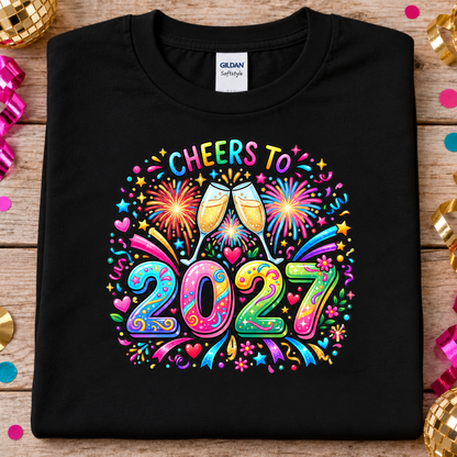 A black Gildan Softstyle t-shirt laid flat on a rustic wooden surface, decorated with festive confetti and gold disco balls. The shirt features a vibrant, colorful graphic on the front with two clinking champagne glasses surrounded by fireworks and the text 'CHEERS TO 2027' in a playful, neon-style font.