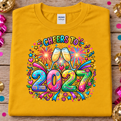 A folded gold colored Gildan Softstyle T-shirt is displayed flat on a rustic wooden surface, decorated with festive confetti and disco ball props. The front of the shirt features a vibrant, colorful graphic that reads 'CHEERS TO 2027' with two clinking champagne glasses, fireworks, and playful party doodles.