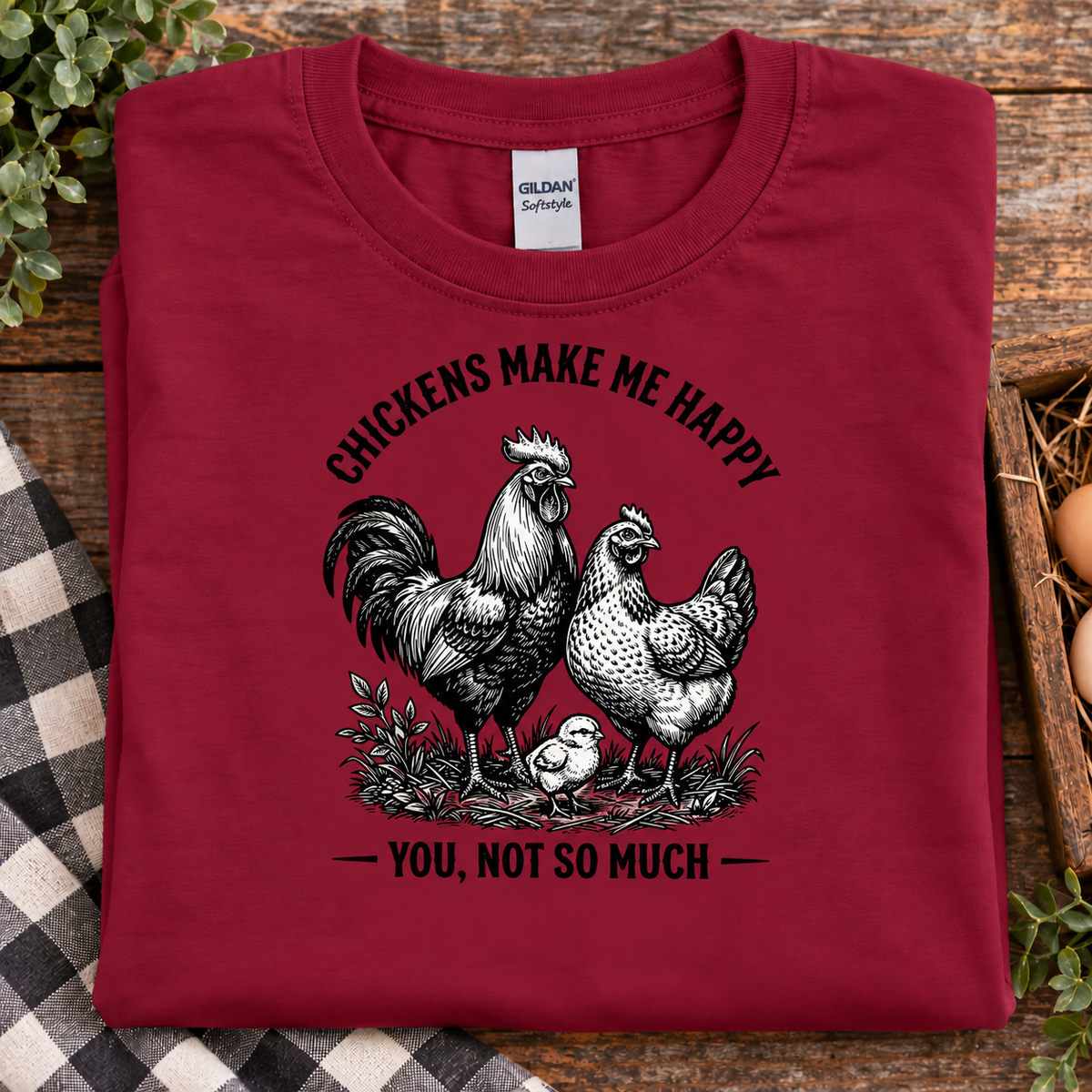 A folded cardinal red Gildan Softstyle t-shirt with a black and white graphic of a rooster, hen, and chick under the text 'CHICKENS MAKE ME HAPPY YOU, NOT SO MUCH', displayed on a rustic wooden background with a plaid cloth.