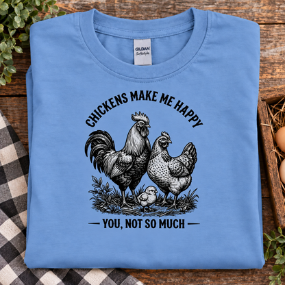 A folded carolina blue Gildan Comfort T-shirt is displayed on a rustic wooden surface with a plaid cloth and greenery. The shirt features a black graphic of a rooster, hen, and chick with the text 'CHICKENS MAKE ME HAPPY YOU, NOT SO MUCH'.