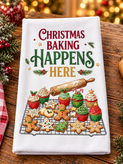 A white kitchen towel with a festive Christmas Baking graphic, displayed on a rustic wooden table with holiday decorations and warm bokeh lights in the background. The graphic reads 'Christmas Baking Happens Here' in red, green, and gold lettering, above an illustration of decorated cupcakes, gingerbread cookies, and a rolling pin.