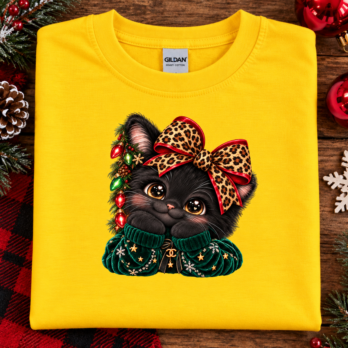 A folded daisy yellow Gildan t-shirt with a graphic of a black kitten wearing a leopard print bow and a green sweater, displayed on a rustic wooden background with Christmas decorations.