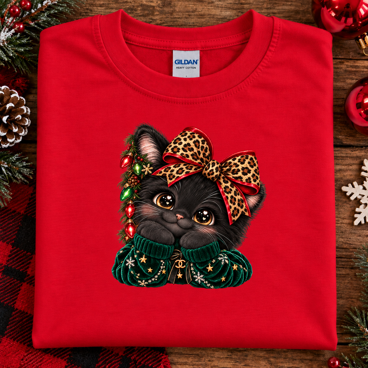 A folded red Gildan sweatshirt with a graphic of a black kitten wearing a leopard-print bow and a green Christmas sweater, surrounded by holiday decorations on a wooden surface.