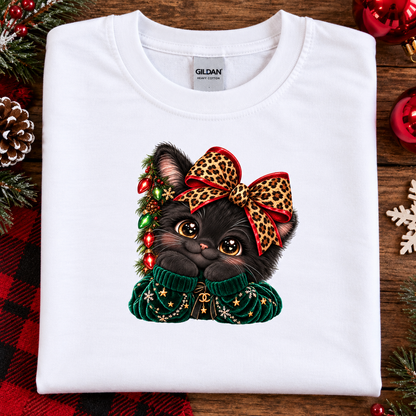A folded white Gildan t-shirt with a Christmas-themed graphic of a black kitten wearing a leopard-print bow and a green knit sweater, displayed on a rustic wooden background with holiday decorations.