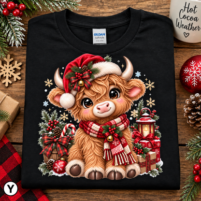 A black Gildan G-1000 t-shirt laid flat on a rustic wooden surface, decorated with a festive Christmas Highland cow graphic. The graphic features a fluffy light brown cow wearing a red Santa hat and a red-and-white plaid scarf, surrounded by holiday decorations like a lantern, candy canes, and ornaments.