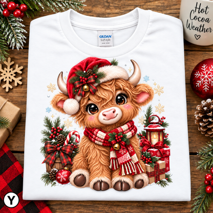 A folded white Gildan t-shirt with a colorful graphic of a cute Highland cow in a red Santa hat and plaid scarf, surrounded by Christmas decorations, displayed on a wooden surface.