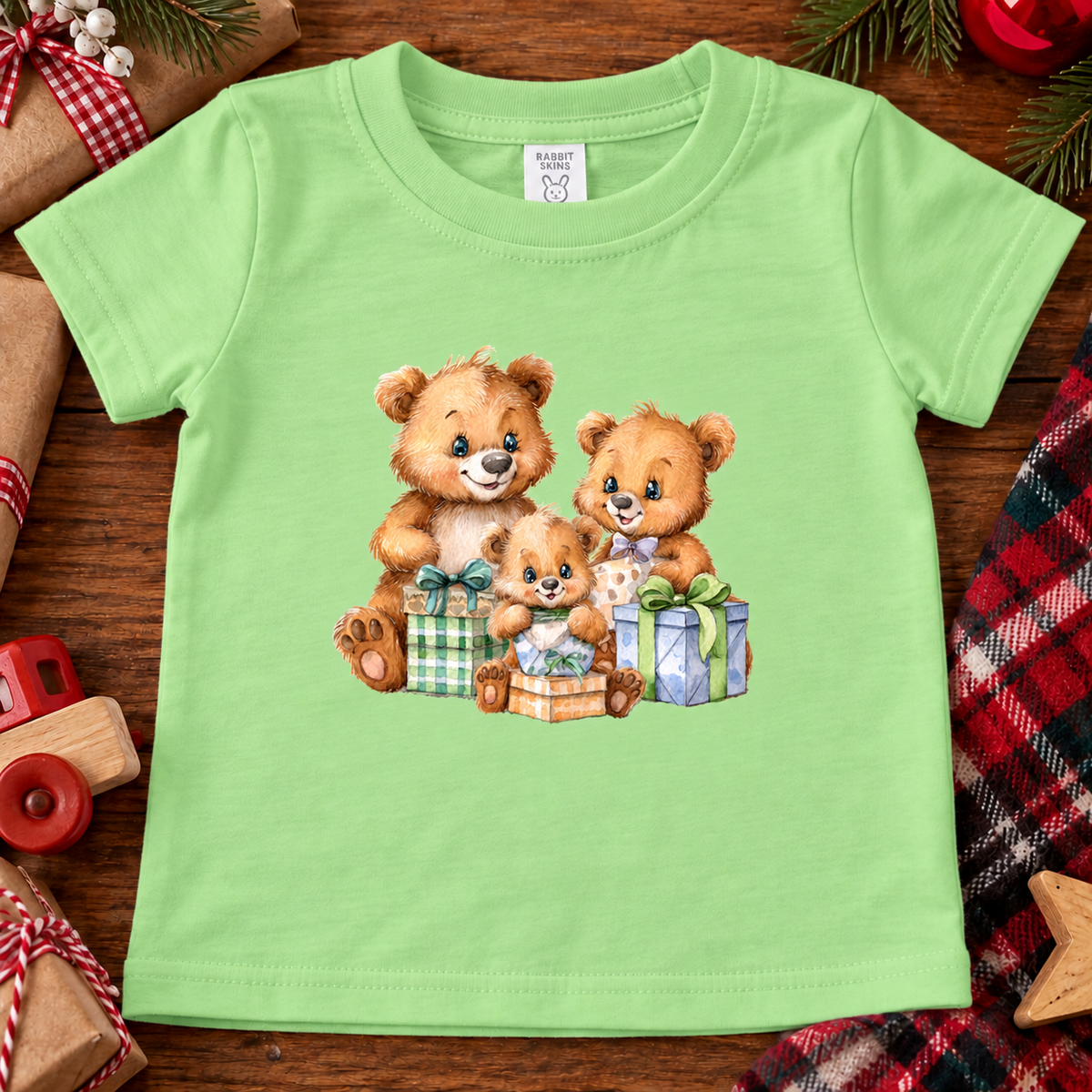 A keylime green short-sleeve baby t-shirt featuring a printed graphic of three cute teddy bears hugging and playing with wrapped Christmas presents. The shirt is displayed flat on a rustic wooden surface surrounded by holiday-themed props like a toy car, plaid fabric, and pine cones.